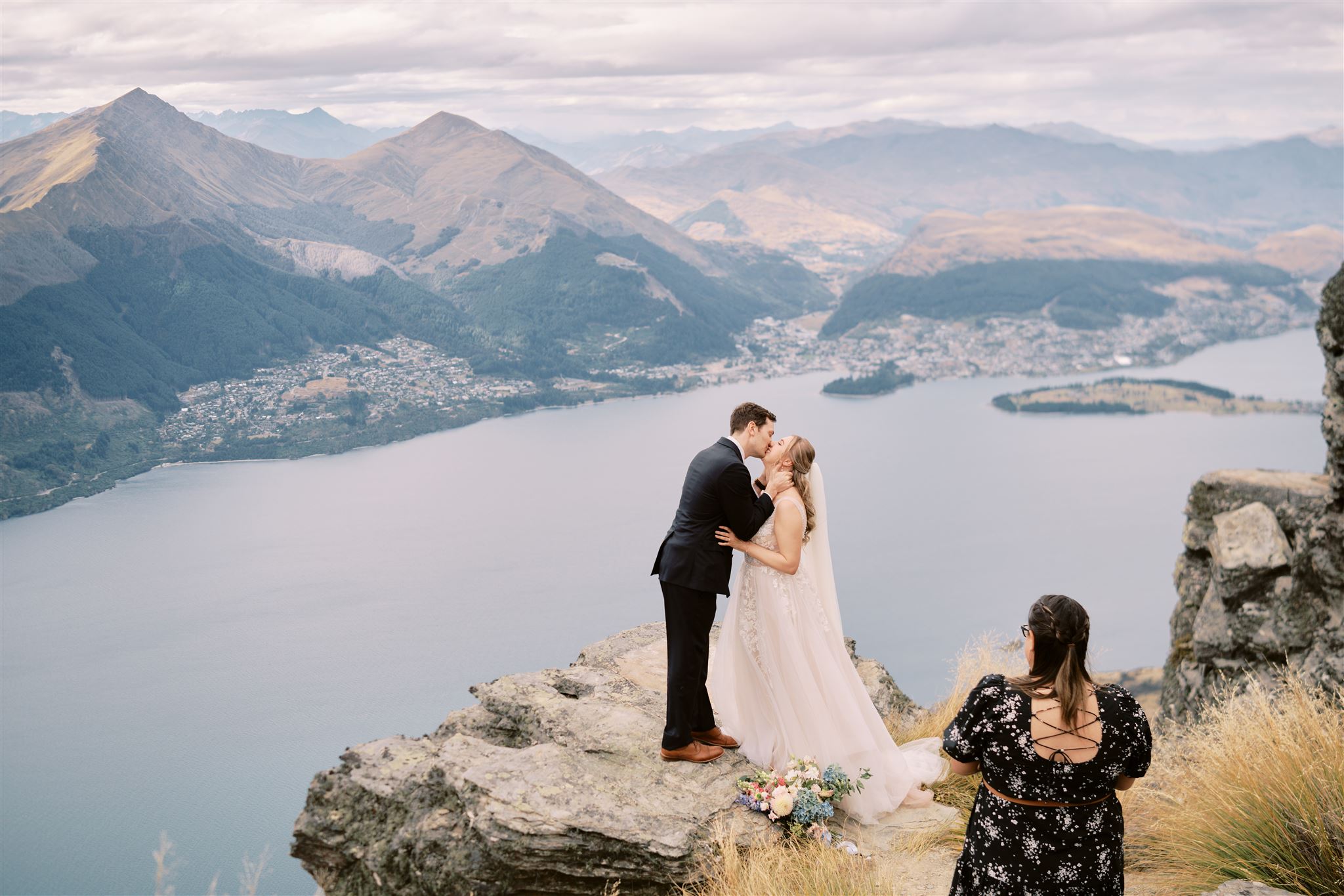 Queenstown New Zealand Heli Wedding Elopement Photographer クイーンズタウン　ニュージーランド　エロープメント 結婚式 | A couple sharing a kiss on a mountain overlook with a lake and hills in the background, while a wedding photographer captures the moment.
